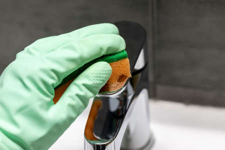 A hand in a mint green rubber glove wipes a chrome metal faucet with an orange sponge. Cleaning in a modern bathroom. Selective focus. Closeup viewの写真素材