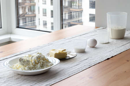 Prepared ingredients: containers with flour, butter, egg, sugar, baking powder, cottage cheese stands on a tablecloth on a wooden table near the window. The process of making curd bagels or croissantsの写真素材