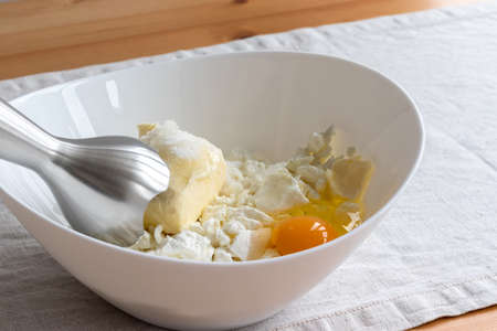 Cottage cheese, butter, egg and sugar in a white bowl - ingredients for mixing with a hand blender on a linen tablecloth on a wooden table. The process of making curd bagels or croissants