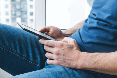 A man in a blue t-shirt and jeans holds an e-reader electronic book in his hands. Reading books at home by the window. Selective focusの写真素材