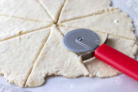 Rolled raw curd dough with bits of cottage cheese cut into eight triangles with a red round pizza knife on it on baking paper closeup. The process of making curd bagels or croissantsの写真素材