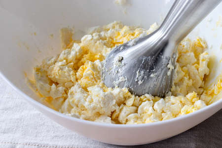 Mixing curd dough in a white bowl with a hand blender. Ingredients - cottage cheese, butter, egg and sugar. The process of making curd bagels or croissantsの写真素材