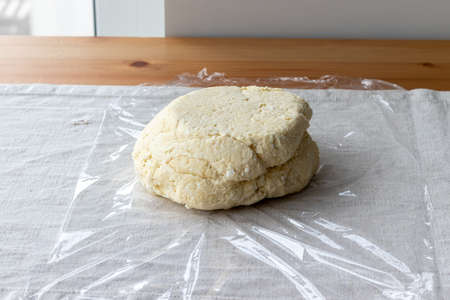 Raw curd dough with bits of cottage cheese on a plastic wrap on a linen tablecloth on a wooden table. The process of making curd bagels or croissantsの写真素材