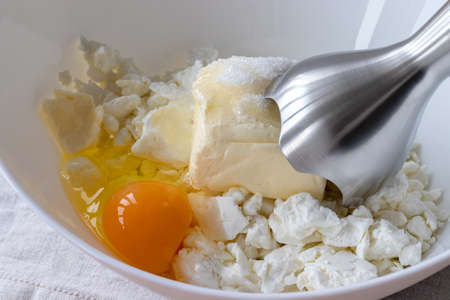 Cottage cheese, butter, egg and sugar in a white bowl - ingredients for mixing with a hand blender. The process of making curd bagels or croissantsの写真素材
