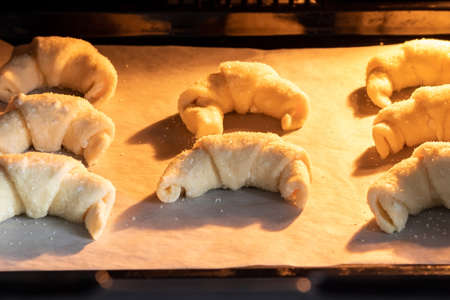 Oven-baked sugar-frosted croissants on a baking sheet covered with parchment paper. Sweet bagels from a cottage cheese dough baked in a hot oven. The process of making curd bagels or croissantsの写真素材