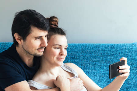 Couple looking at the mobile phone hugging and smiling. Man and woman, family of two, call friends or parents online, take selfies, watch positive videos during quarantineの写真素材