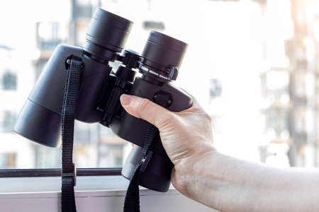 Hand holding black binoculars against a blurred background of a window with a view of the neighboring house for observing neighbors, environment or nature. Selective focus. Closeup viewの写真素材
