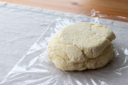 Raw curd dough with bits of cottage cheese on a plastic wrap on a linen tablecloth on a wooden table. The process of making curd bagels or croissants. Closeup viewの写真素材