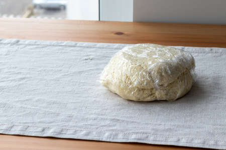 Raw curd dough with bits of cottage cheese is wrapped in plastic wrap on a linen tablecloth on a wooden table. The process of making curd bagels or croissantsの写真素材
