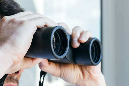 Man looking through binoculars at the window with a view of the neighboring house for observing neighbors, environment or nature during quarantine. Selective focus. Closeup viewの写真素材