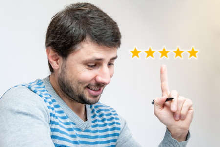 Handsome smiling bearded man with a pen in his hand pointing his index finger upwards to five luminous orange stars. Customer gives the highest rating to the product or application in the online storeの写真素材
