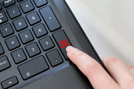 Keyboard corner of a black laptop with a female finger clicking on a fake news button. The concept of spreading false news, messages on the Internetの写真素材