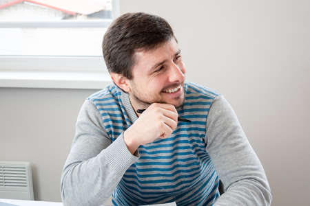 Handsome smiling man in a striped sweater sits in an office with a pen in his hand and looking awayの写真素材