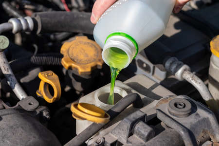 Hand with bottle pouring antifreeze coolant into the expansion tank. Dusty details of a flat-four (boxer) car engine compartment under the open hood. Closeup view on a sunny dayの写真素材