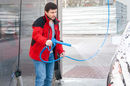 A man in a red jacket covers a black car with foam at a self-service car washの写真素材