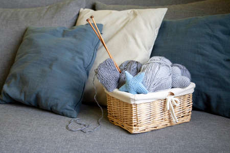 Gray balls of wool and knitting needles in a wicker basket stand on a gray sofa with pillowsの写真素材