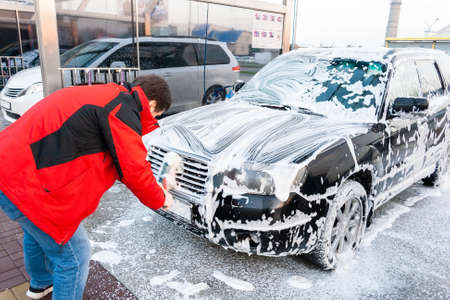 A man in a red jacket wipes a foam-covered black car with a brush at a self-service car wash. Front viewの写真素材