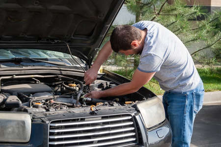 A bearded man leaned over the open hood of a black car with a screwdriver during the repair process. Diagnosis and replacement of failed car parts yourselfの写真素材