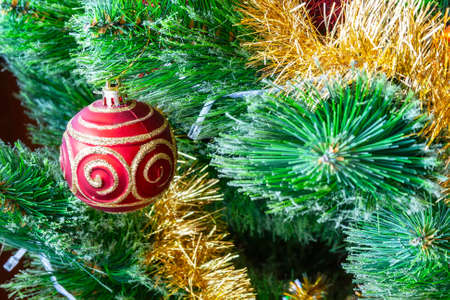Red ball with gold glitter patterns hangs on a green artificial Christmas tree among the golden garlands for a family holiday. New Year's Eve home decorationの写真素材