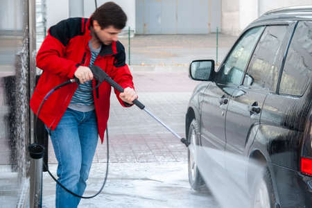 A man in a red jacket washes a black car with a strong jet of water at a self-service car washの写真素材