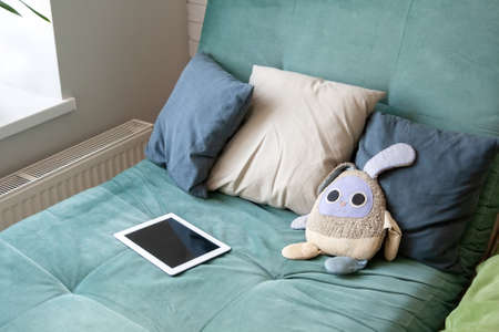 Gray pillows, a white tablet and a toy bunny lying on a soft, comfortable, cozy green couch near the window. Beautiful modern living room interiorの写真素材