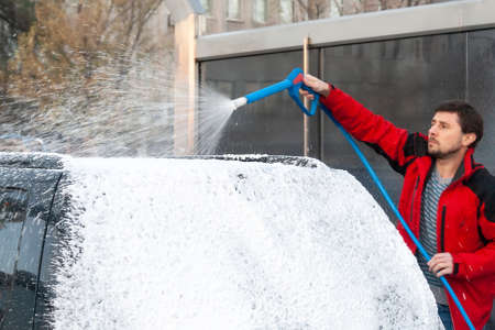 A man in a red jacket covers a black car with foam at a self-service car wash. Back viewの写真素材