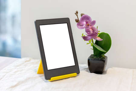 Modern gray electronic book or tablet with a blank mockup screen on a yellow stand next to an artificial pink orchid on the kitchen table with a linen tablecloth by the windowの写真素材