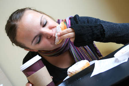 A beautiful woman in a striped scarf, dark gray sweater and mittens holds a cardboard cup of coffee in her hand and bites a donut. There is a tray and a second white-glazed donut on the tableの写真素材