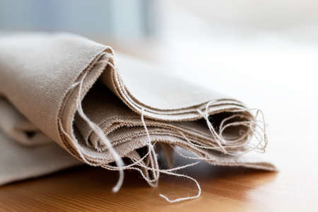 Roll of natural linen fabric with protruding threads on a wooden table. Selective focus. Closeup view. Blurred background