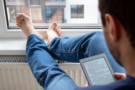 Over the shoulder view of relaxing man in a blue jeans reading an electronic book on digital device by the window. Focus on bare feet lying on a windowsill in a bright modern apartmentの写真素材