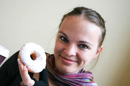 Smiling beautiful woman in a multi-colored striped scarf, dark gray sweater and mittens is holding a donut in white glazeの写真素材