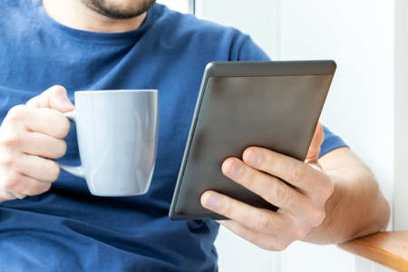 A man in a blue t-shirt holds a gray e-reader electronic book in one hand and a cup in the other. Reading a book or news on a tablet at breakfast. Selective focusの写真素材