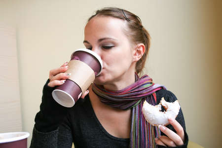 Beautiful woman in a multi-colored striped scarf, dark gray sweater and mittens is drinking coffee from a cardboard cup and holding a bitten donut in her other handの写真素材