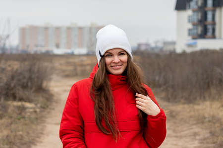 Smiling beautiful young woman with long hair in a white knitted hat and a red jacket with a hood on the street on a cloudy day against a blurred background. Girl without makeup, natural beautyの写真素材