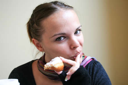 Beautiful woman in a multi-colored striped scarf, dark gray sweater and mittens licks her finger, holding a donut in her hands, and smiling slyly looks into the cameraの写真素材
