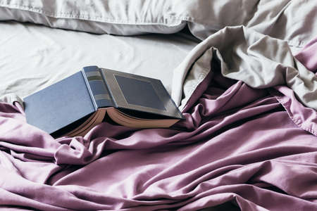 Open blue book on the bed with a purple bedspread and gray pillows. Fascinating book in soft bedding