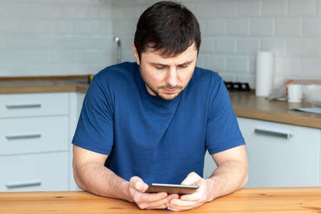 Handsome bearded man in a blue T-shirt sits behind a wooden table and reads an electronic book against a blurred background of a modern kitchenの写真素材