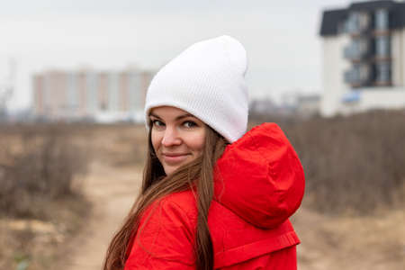 Smiling young woman with long hair in white knitted hat and red jacket with hood looking over her shoulder on the street on cloudy day against blurred background. Female without makeup, natural beautyの写真素材