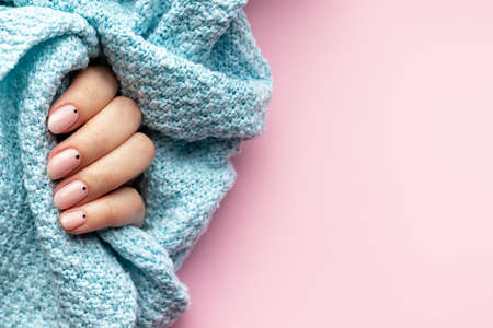 Female hand in a blue knitted sweater fabric with trendy beautiful manicure - pink nude nails with black small dots on a pink background with copy spaceの写真素材