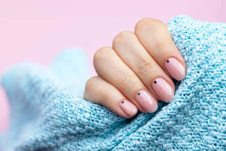 Female hand in a blue knitted sweater fabric with trendy beautiful manicure - pink nude nails with black small dots on a pink background with copy spaceの写真素材
