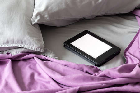 A modern black e-reader electronic book with a blank screen on a bed with two gray pillows and a...