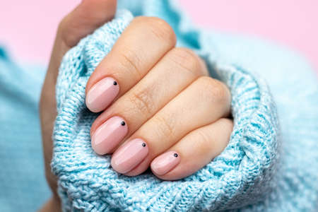 Female hand in a blue knitted sweater fabric with trendy beautiful manicure - pink nude nails with black small dots on a pink background. Selective focus. Closeup viewの写真素材
