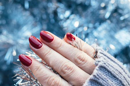 Female hand in a gray knitted sweater with a beautiful glossy manicure - burgundy, dark red, cherry color nails on background of silver Christmas tinsel garland. Selective focus. Blurred backgroundの写真素材