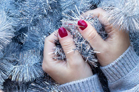 Female hands in a gray knitted sweater with a beautiful glossy manicure - burgundy, dark red, cherry color nails on background of silver Christmas tinsel garlandの写真素材