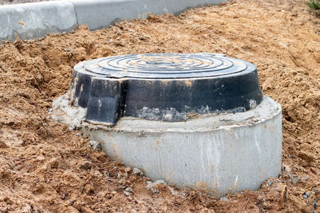 A black cast-iron sewer hatch on a concrete base, located on a mound of damp ground for access to...