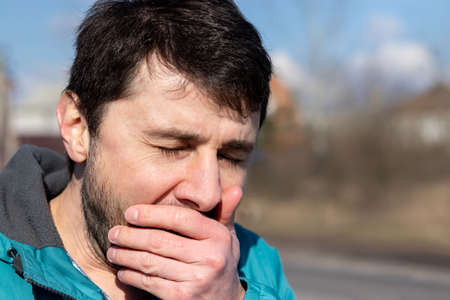 Coughing, sneezing or yawning squinting bearded man, covering his mouth with his hand on the street on a sunny day. Sick or tired man. Closeup view. Blurred backgroundの写真素材