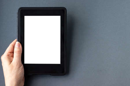 A modern black electronic book with a white blank empty screen in female hand on a gray paper background. Mockup tablet closeup with copy spaceの写真素材