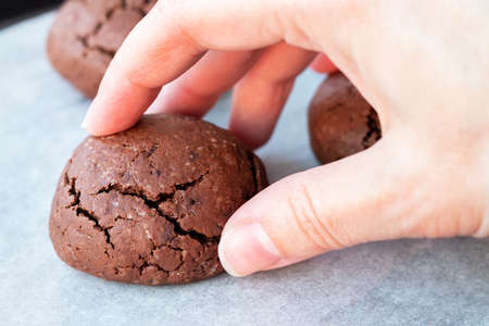 Hand takes baked cracked round chocolate cookie from a baking sheet with parchment paper. Tea or...