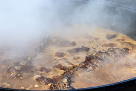 Cooking real Uzbek pilaf. Stewing lamb ribs in a huge cauldron with lots of steam outside during a...