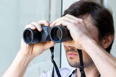 Handsome bearded man looking through binoculars at the window with a view of the neighboring house...